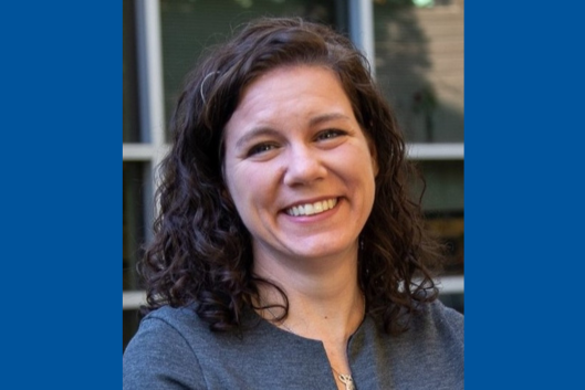 Pictured is Dr. Karen Miga, a woman with shoulder-length, curly dark brown hair is smiling at the camera. She is wearing a dark gray top and a delicate necklace. The background is slightly blurred, featuring a building with large windows. Natural lighting highlights her face.